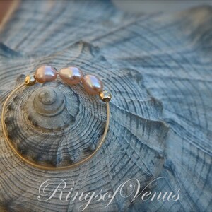 May include: A gold ring with three pink pearls on a seashell background. The ring is made of thin wire and the pearls are small and round. The text "Rings of Venus" is visible in the bottom right corner of the image.