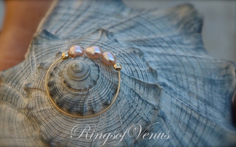 May include: A gold ring with three pink pearls on a seashell background. The ring is made of thin wire and the pearls are small and round. The text "Rings of Venus" is visible in the bottom right corner of the image.