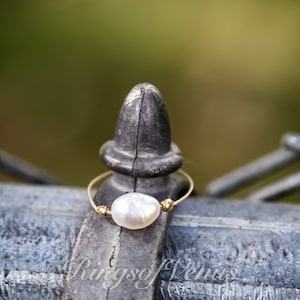 May include: A delicate gold ring featuring a single white pearl. The ring is resting on a metal fence post.