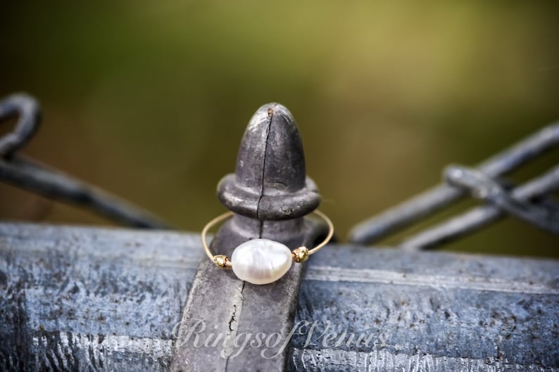 May include: A delicate gold ring featuring a single white pearl. The ring is resting on a metal fence post.