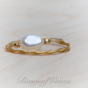 May include: A gold braided ring with a white pearl accent. The ring is on a light wood surface.