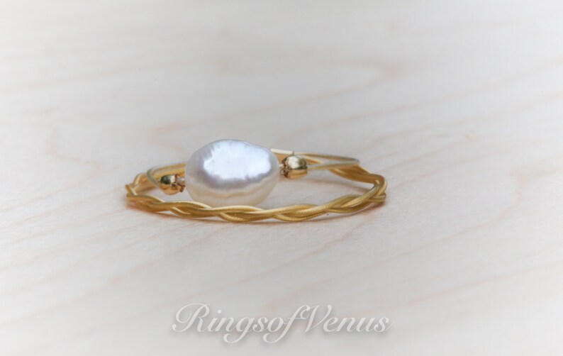 May include: A gold braided ring with a white pearl accent. The ring is on a light wood surface.