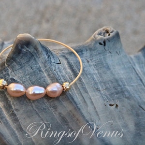 May include: A gold bangle bracelet with three pink pearls and gold beads. The bracelet is on a piece of driftwood with the text "Rings of Venus" in the bottom right corner.