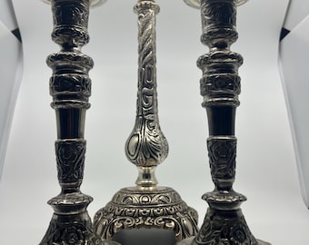May include: Set of three silver candle holders with ornate designs. The center candle holder is taller and has a more elaborate design than the two side candle holders.