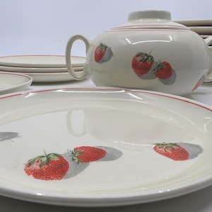 May include: Cream-colored ceramic dinnerware set with a red trim and strawberry design. Includes a teapot, plates, and bowls. The strawberries have a gray shadow. The teapot has a lid and two handles.