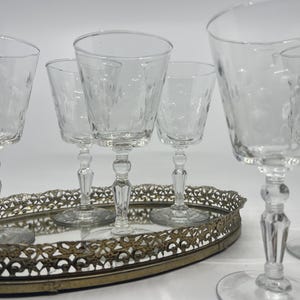 May include: A collection of clear, etched glass wine glasses with ornate stems, arranged on a decorative, oval-shaped metal tray. The glasses have a delicate floral pattern and are set against a white background.