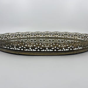 May include: An oval, antique-style vanity tray with a mirrored base and a decorative, openwork metal rim. The rim has a detailed, floral-inspired design and a tarnished gold finish. The tray is likely made of metal and glass.