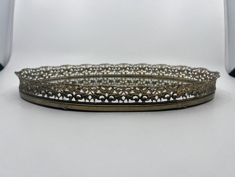 May include: An oval, antique-style vanity tray with a mirrored base and a decorative, openwork metal rim. The rim has a detailed, floral-inspired design and a tarnished gold finish. The tray is likely made of metal and glass.