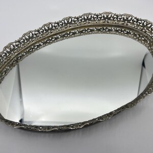 May include: Oval silver-toned vanity tray with a mirrored surface. The tray features an ornate, openwork filigree border. The mirror reflects the surrounding environment. The tray is likely vintage and suitable for displaying jewelry or perfumes.