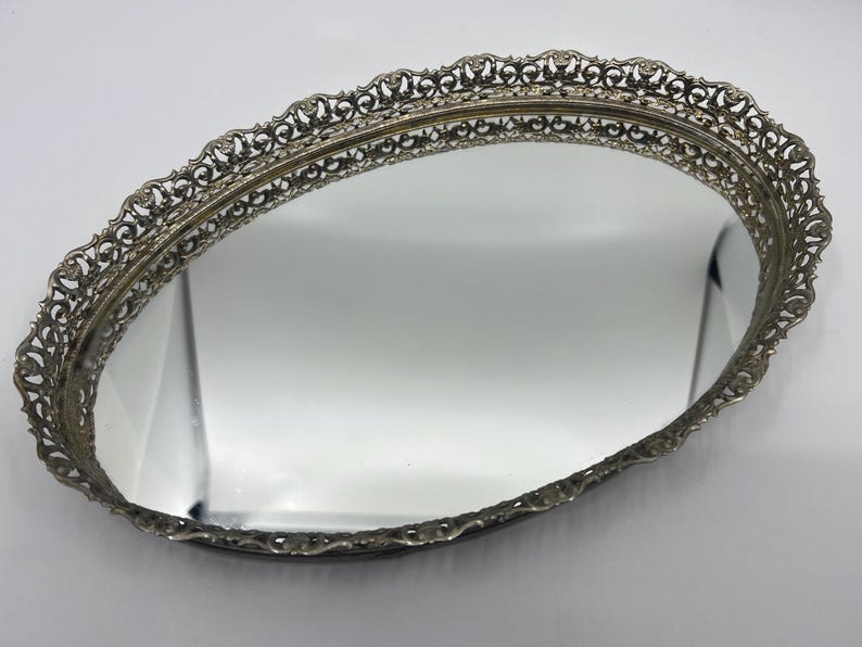 May include: Oval silver-toned vanity tray with a mirrored surface. The tray features an ornate, openwork filigree border. The mirror reflects the surrounding environment. The tray is likely vintage and suitable for displaying jewelry or perfumes.