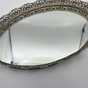 May include: Oval vanity mirror with a decorative filigree metal frame. The mirror has a reflective surface and a detailed, ornate border. The frame is a silver color, and the mirror is set against a white background.