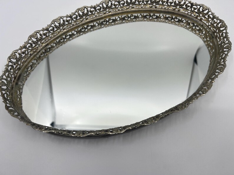 May include: Oval vanity mirror with a decorative filigree metal frame. The mirror has a reflective surface and a detailed, ornate border. The frame is a silver color, and the mirror is set against a white background.