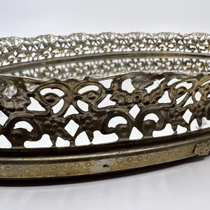 May include: An oval, vintage vanity tray with a mirrored base and ornate, openwork metal filigree border. The metal has a tarnished, antique gold finish. The tray's design features floral and scroll motifs, adding a touch of elegance.