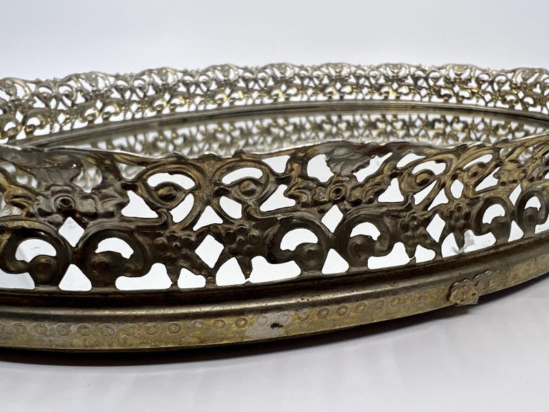 May include: An oval, vintage vanity tray with a mirrored base and ornate, openwork metal filigree border. The metal has a tarnished, antique gold finish. The tray's design features floral and scroll motifs, adding a touch of elegance.