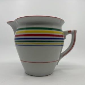 May include: White ceramic pitcher with a red rim and handle accents. The pitcher features horizontal stripes in green, yellow, red, blue, and a thin red stripe near the base. It has a spout for pouring.