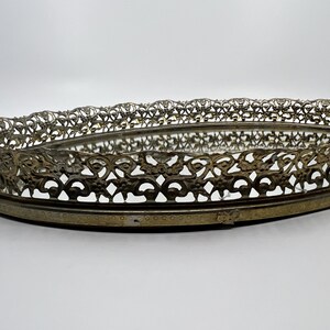 May include: An oval-shaped vintage vanity tray with a mirrored base and ornate, openwork metal filigree edging. The metal has a tarnished, antique brass finish. The tray is set against a plain white background.