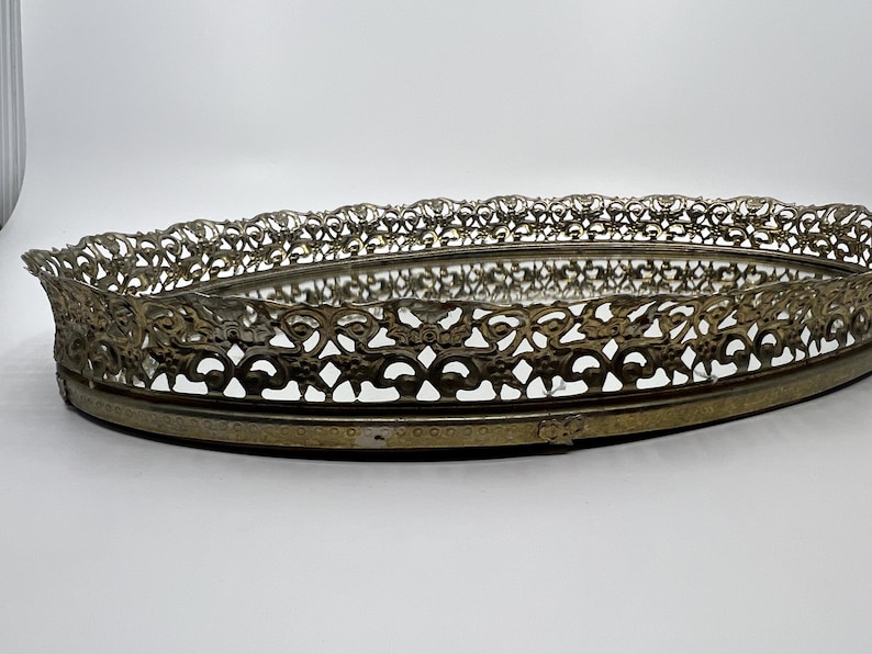 May include: An oval-shaped vintage vanity tray with a mirrored base and ornate, openwork metal filigree edging. The metal has a tarnished, antique brass finish. The tray is set against a plain white background.