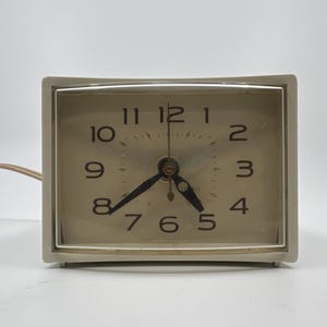 May include: A vintage, rectangular alarm clock with a cream-colored face and a light beige frame. The clock face features black numerals and hands, with a second hand. A power cord extends from the back. The clock is a classic design.