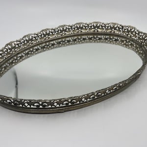 May include: Oval vanity tray with a mirrored surface and an ornate, filigree-style metal frame. The frame has a scalloped edge and a tarnished silver finish, creating a vintage aesthetic. The tray is empty and reflects the surrounding environment.