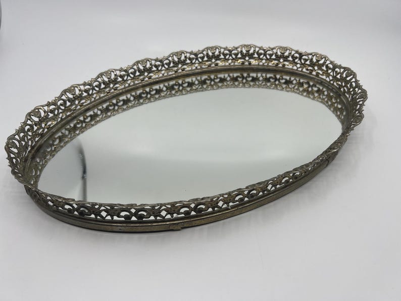 May include: Oval vanity tray with a mirrored surface and an ornate, filigree-style metal frame. The frame has a scalloped edge and a tarnished silver finish, creating a vintage aesthetic. The tray is empty and reflects the surrounding environment.