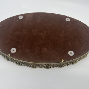 May include: An oval-shaped vanity tray with a brown velvet interior and an ornate, filigree-style metal frame. The tray has small, round feet and is designed for displaying jewelry or perfumes.
