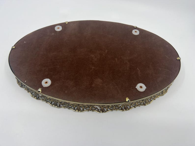 May include: An oval-shaped vanity tray with a brown velvet interior and an ornate, filigree-style metal frame. The tray has small, round feet and is designed for displaying jewelry or perfumes.