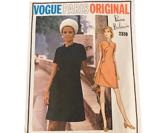 May include: A vintage Vogue Paris Original sewing pattern featuring a woman in a black dress and a fashion illustration of a peach-colored dress. The pattern is labeled "Pierre Balmain 2338."