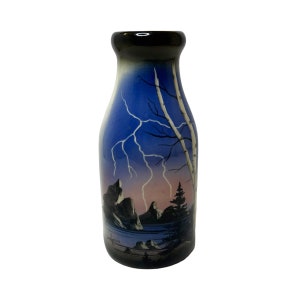 May include: A blue and pink ceramic vase with a black rim and a painted scene of mountains, trees, and lightning. The vase is shaped like a milk bottle and has a signature at the bottom.