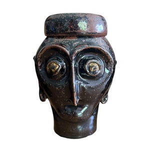 May include: A dark brown ceramic jar with a lid, shaped like a stylized human head. The jar has large, round eyes with gold accents, a long nose, and a small mouth. The surface has a glossy finish with speckled details.