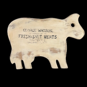 May include: A white wooden cow-shaped cutting board with a distressed finish. The board features a black painted sign that reads "George Watson, Dealer in Fresh & Salt Meats, Poultry & C., Binghamton, N.Y."