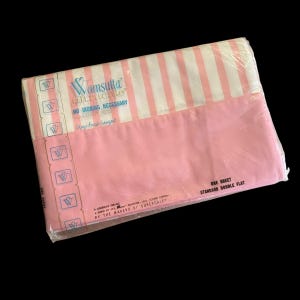 Könnte beinhalten: Ein verpacktes Spannbettlaken in Rosa mit weißen vertikalen Streifen. Auf der Oberseite der Bettlakenverpackung ist der Markenname "Vamsutta ultracale" in Blau und Rosa aufgedruckt. Die Verpackung ist mit "One Sheet Standard Double Flat" beschriftet.