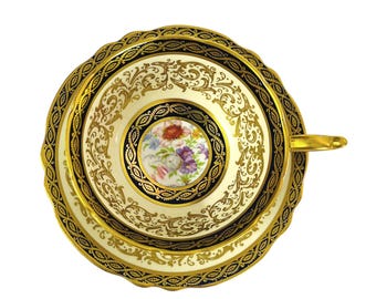 May include: Overhead view of a vintage teacup and saucer set. The ivory-colored cup and saucer feature intricate gold filigree and a black border. The center of the cup displays a floral design with red, purple, and white flowers.
