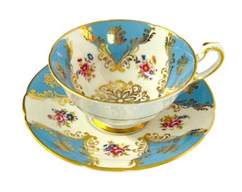 May include: A vintage teacup and saucer set. The teacup and saucer feature a light blue and white design with gold trim. Floral patterns in pink, blue, and yellow adorn the cup and saucer. The teacup has a delicate handle.
