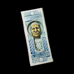 May include: A rectangular ceramic plaque featuring a central relief sculpture of a person's face with a light-colored head covering. The plaque is decorated with intricate blue and white geometric patterns and a circular frame around the face.