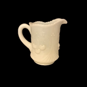 May include: White milk glass jug with a raised design of cherries and leaves. The jug has a handle and a rounded spout.