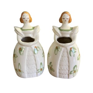 May include: Two ceramic figurines of women in dresses. The figurines have orange hair, green accents, and white dresses decorated with green floral patterns. The figurines have open mouths and closed eyes, and are likely decorative vases or planters.