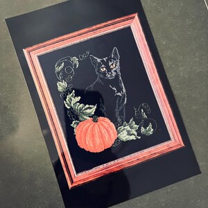 May include: A black cat with green eyes sits in front of a pumpkin and green vines. The image is framed in a dark wood frame.
