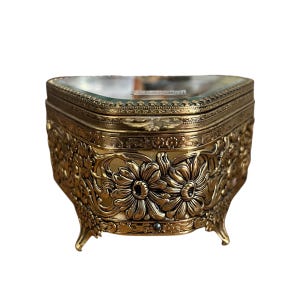 May include: Ornate gold-colored jewelry box with a hinged lid and floral detailing. The box has a rectangular shape with rounded corners and sits on four small, curved feet. The lid has a clear glass top, and the box is made of metal.