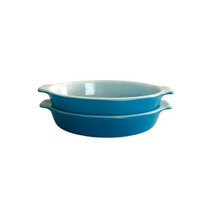 May include: Two oval-shaped, turquoise ceramic baking dishes stacked on top of each other. Each dish has a white interior and small handles on either side. The dishes are set against a white background.