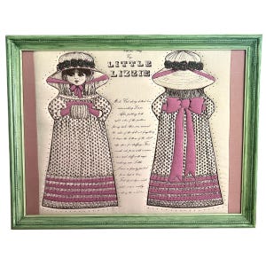 May include: Framed vintage artwork featuring two illustrations of a doll named "Little Lizzie." The doll is depicted in a long dress with a floral pattern and a large hat. The artwork is framed in a green wooden frame with a pink mat.