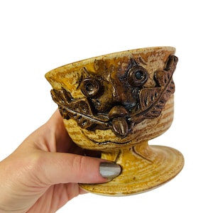 May include: A handmade ceramic owl-faced bowl with a pedestal base. The bowl features a textured, earthy color palette of brown, tan, and cream. The owl's face is detailed with circular eyes, a beak, and leaf-like accents.