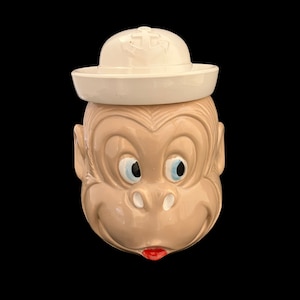 May include: A ceramic monkey cookie jar with a white sailor hat. The monkey has a brown face with blue eyes and a red mouth. The hat has an anchor design on the top.