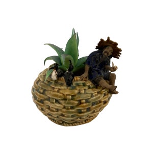 May include: A ceramic planter shaped like a woven basket with a small green succulent plant and a figurine of a man wearing a straw hat sitting on the edge.