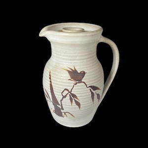 May include: Cream-coloured ceramic jug with a brown bird and branch design. The jug has a handle, a spout, and a lid. The bird is perched on a branch with leaves. The jug is likely used for serving beverages.