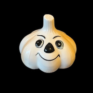 May include: A white ceramic garlic clove with a painted face. The garlic has black eyes, a black nose, and a smiling mouth.