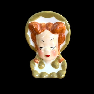 May include: A vintage ceramic wall pocket in the shape of a woman's face. The woman has red hair, closed eyes, and red lips. The wall pocket is primarily white, with green and orange accents. The design is reminiscent of mid-century decor.