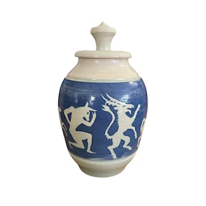 May include: A ceramic jar with a lid. The jar has a blue body with white figures and a cream-coloured top. The jar is rounded with a lid and a decorative finial. The white figures appear to be mythical creatures.