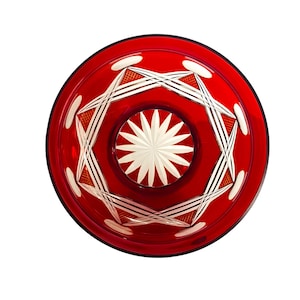 May include: A top-down view of a red glass bowl with a starburst design in the center. The bowl has a black rim and white and red geometric patterns. The center of the bowl features a white star-shaped design.