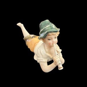 May include: A porcelain figurine of a young person lying down and playing a flute. The figurine wears a green hat, white shirt, and yellow shorts. The figure is on a black background.