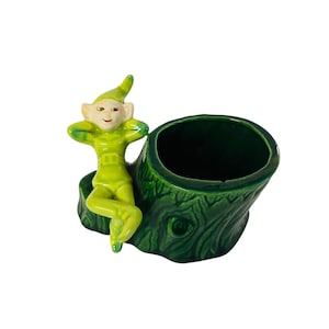 May include: A ceramic planter featuring a green elf figure leaning against a dark green tree stump. The elf is wearing a green outfit and a pointed hat. The planter has a dark green interior and a textured exterior.